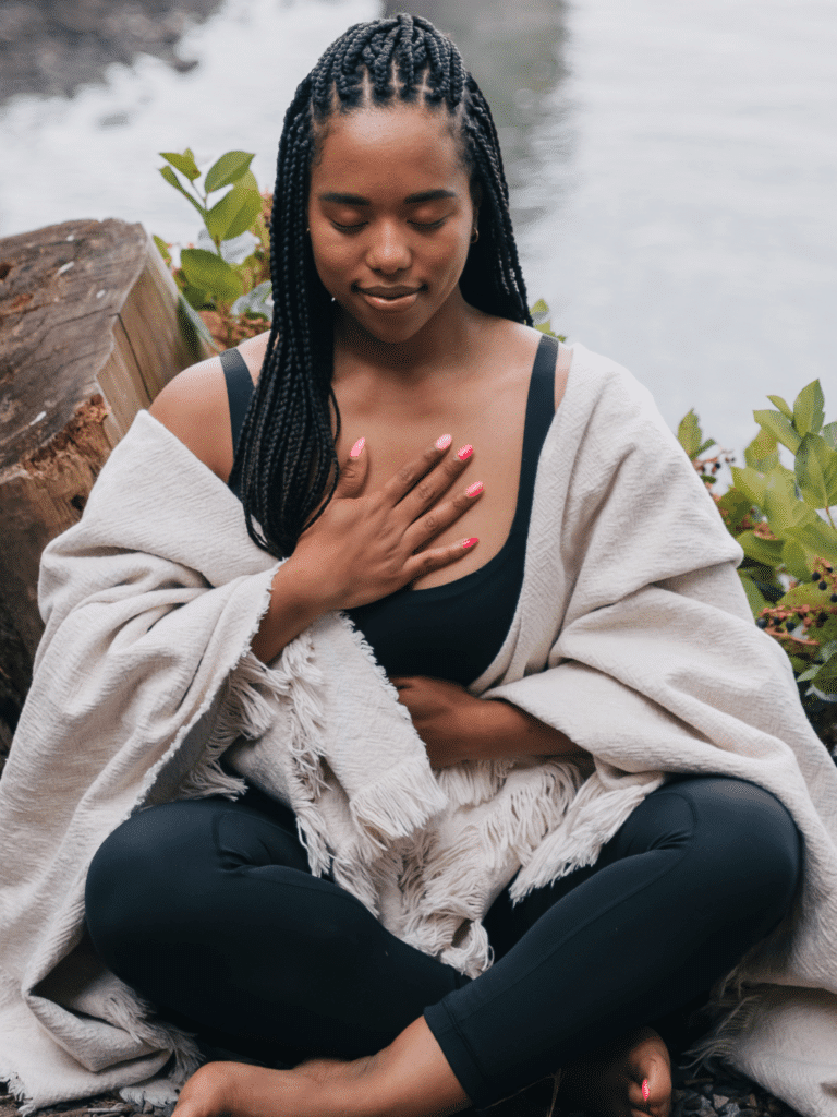 woman meditates next to water, resting one hand on heart and one on stomach.